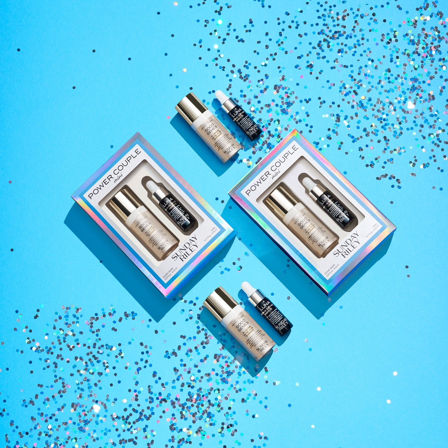 Four pairs of Luna and Good Genes, two in their iridescent Power Couple box, and two without, laid flat in a diagonal square on a blue background with glitter on the top right and bottom lefthand corners