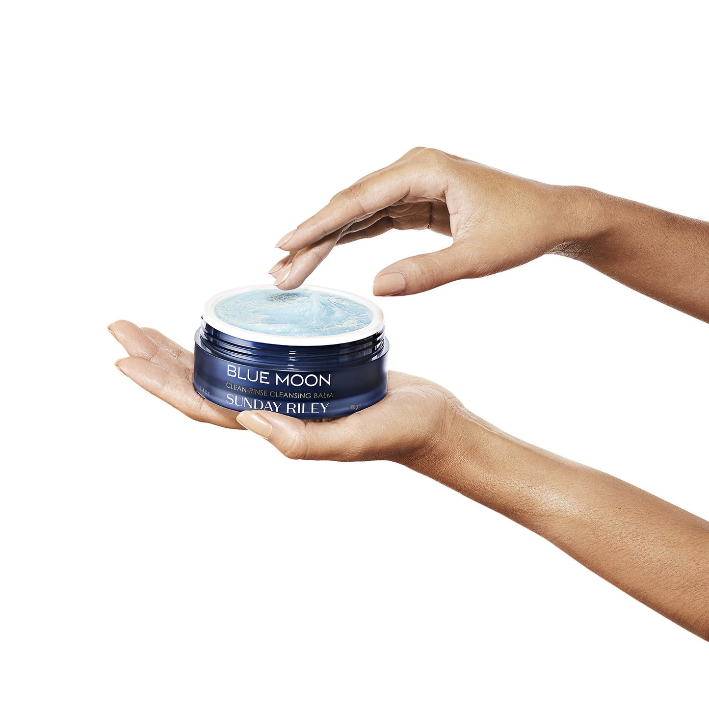 Hand holding Blue Moon Cleansing Balm open jar with opposite hand touching light blue sugar like balm.