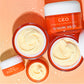 C.E.O. Vitamin C Cream 15g jars and 50g jars lay over peach background with water drops around the jars